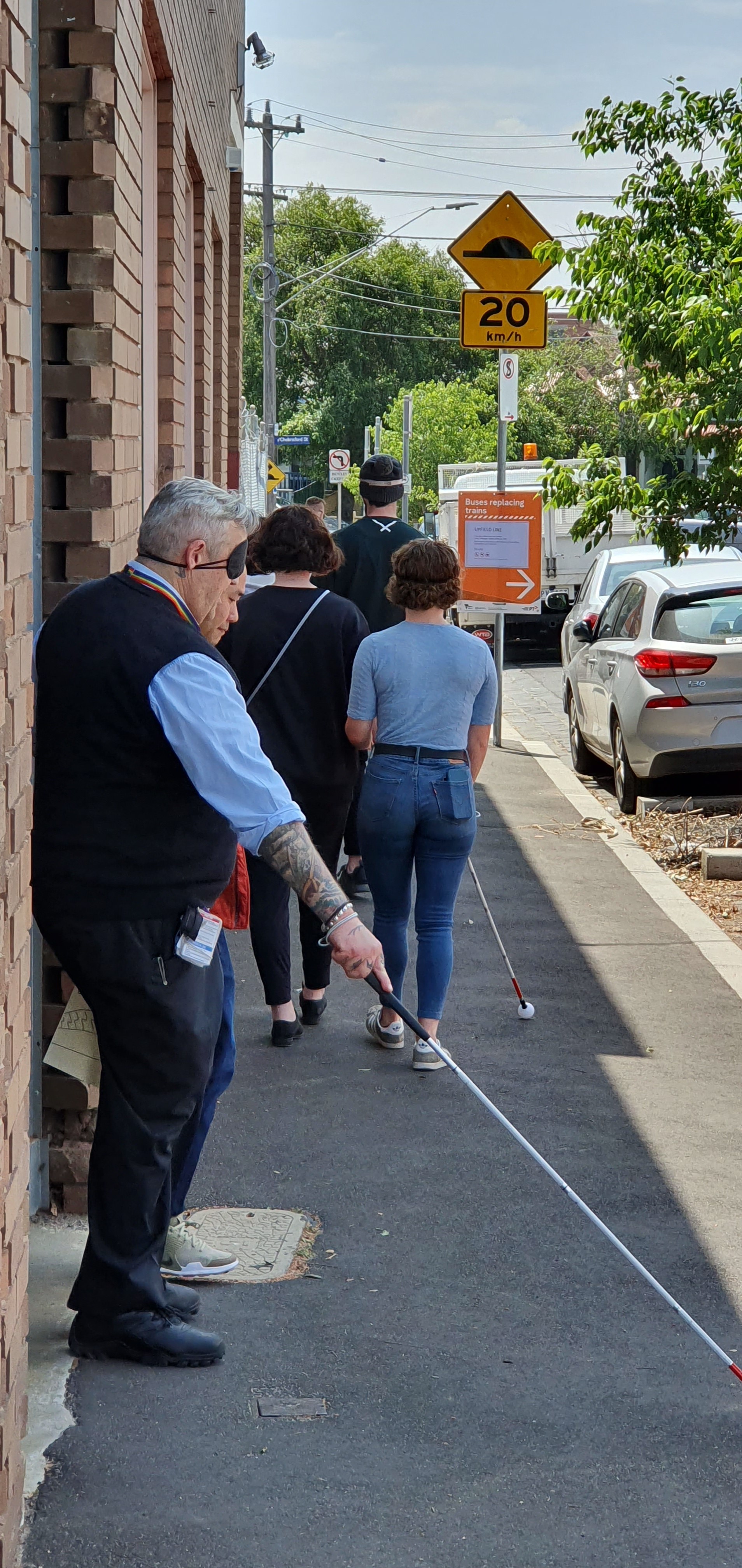 A blindfolded Metro Trains staff member uses a cane to navigate the pavement under the guide of another staff member.