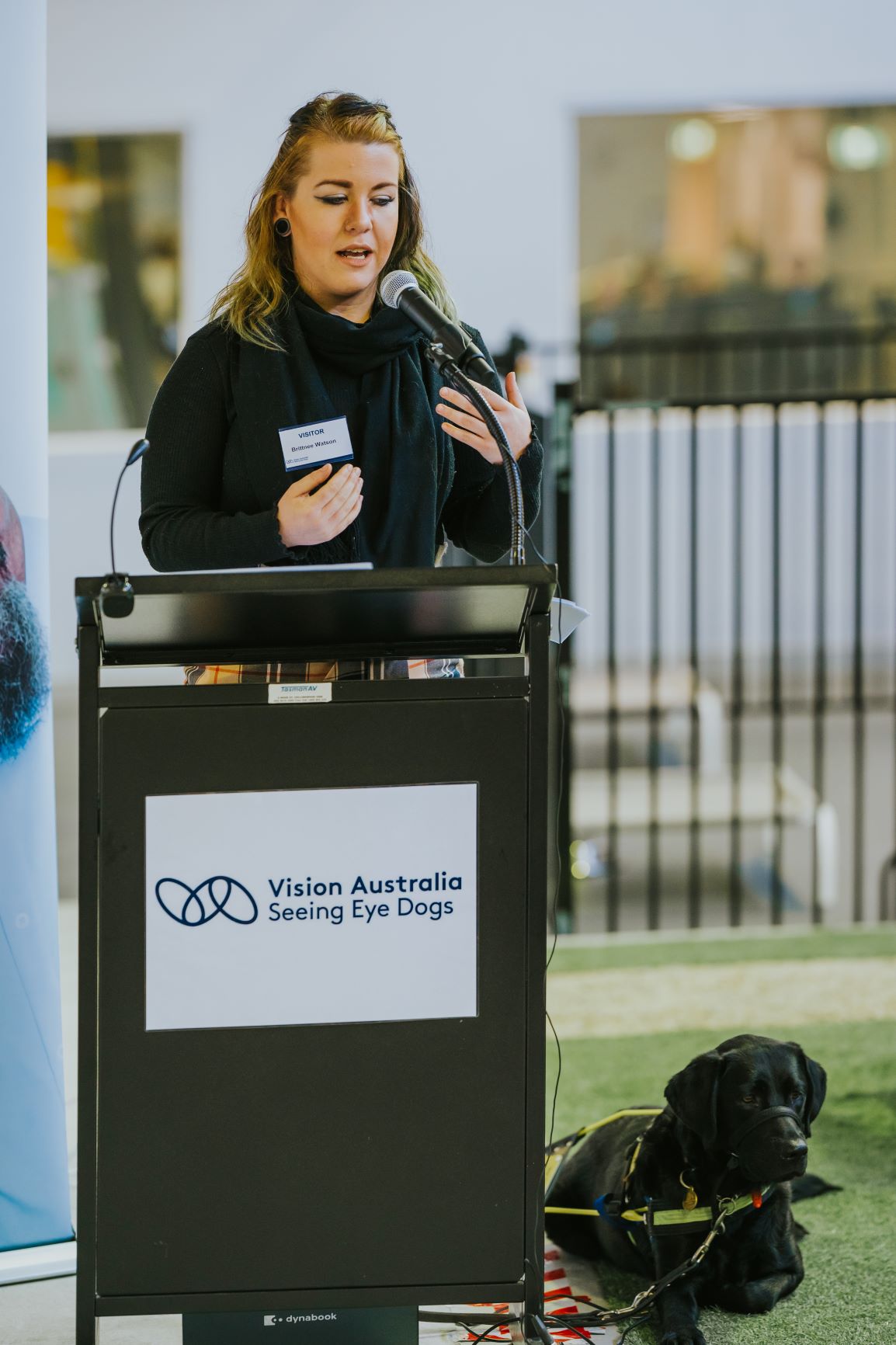 Client Britnee speaking at a lectern during the ceremony with her Seeing Eye Dog Kuma by her side.